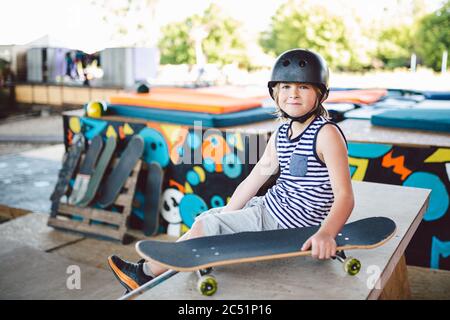 Ein hübscher kaukasischer Junge in einem Helm sitzt auf einer Rampe in einem Skatepark. Ein Skateboarder ruht zwischen den Tricks. Athlet Kinderpause für ein Skateboard Stockfoto