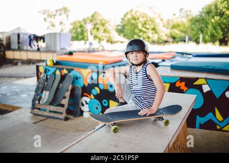 Ein hübscher kaukasischer Junge in einem Helm sitzt auf einer Rampe in einem Skatepark. Ein Skateboarder ruht zwischen den Tricks. Athlet Kinderpause für ein Skateboard Stockfoto