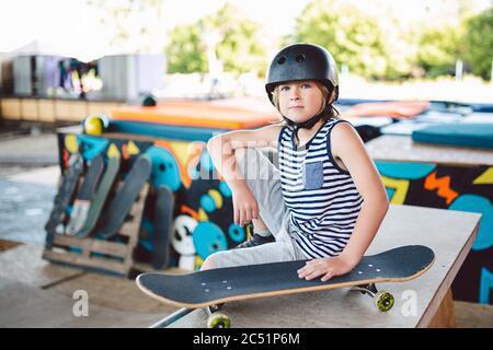 Junge sitzt auf Skatepark, Kamera schauen. Kind ruht mit Skateboard im Skatepark. Kind sitzt auf Rampe ruht nach dem Training auf Skateboard. Trendy Stockfoto