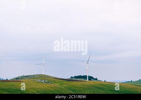 Zwei riesige Windturbinen zwischen den grünen Hügeln gegen den bewölkten Himmel. Stockfoto