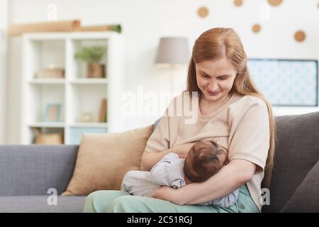 Porträt von glücklich reifen Mutter Stillen Baby während auf der Couch im Haus Innenraum sitzen, kopieren Raum Stockfoto