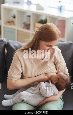 Vertikales Porträt von glücklich reifen Mutter Stillen Baby während auf der Couch im Haus Innenraum sitzen, kopieren Raum Stockfoto