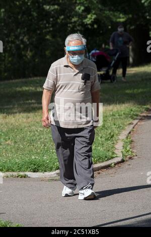 Ein älterer asiatischer Amerikaner, wahrscheinlich Chinese American, führt im Kissena Park mit einer chirurgischen Maske und einem Gesichtsschutz aus Kunststoff durch. Stockfoto