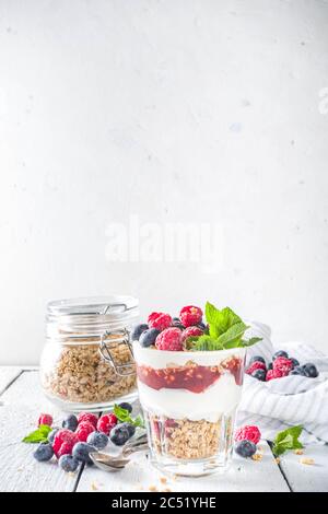 Joghurt parfafait mit Müsli und Beeren. Süßes und gesundes Frühstücksdessert im Glas mit Müsli, Joghurt, Heidelbeeren und Himbeeren. Weißes Holz Stockfoto