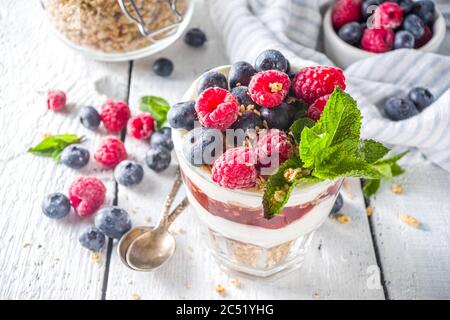 Joghurt parfafait mit Müsli und Beeren. Süßes und gesundes Frühstücksdessert im Glas mit Müsli, Joghurt, Heidelbeeren und Himbeeren. Weißes Holz Stockfoto