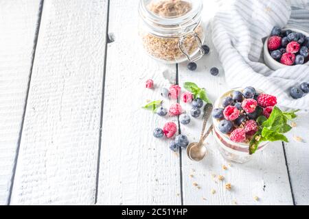 Joghurt parfafait mit Müsli und Beeren. Süßes und gesundes Frühstücksdessert im Glas mit Müsli, Joghurt, Heidelbeeren und Himbeeren. Weißes Holz Stockfoto