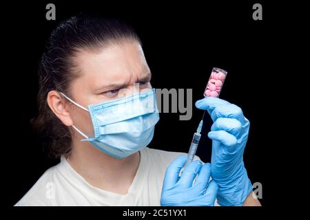 Ein Arzt in einer medizinischen Maske und sterilen blauen Handschuhen hält in seinen Händen eine Spritze mit einer Nadel und eine Glasflasche mit rosa Tabletten, Illusion Stockfoto