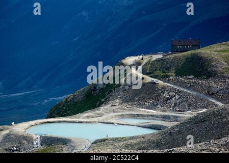 Blick auf kleinen See mit blauem Wasser in Bergregion. Silhouette des Wanderers, der am Ufer läuft. Felsige Bergstraße mit Autos und Touristenherbergen auf dem Hügel. Konzept von Natur und Hochland Stockfoto