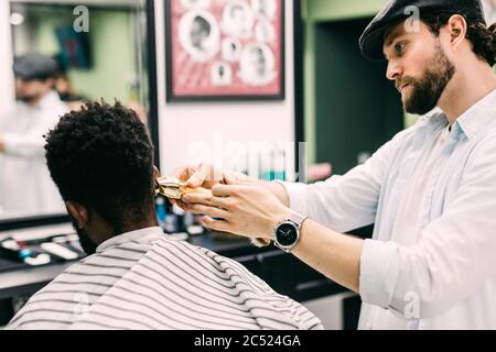 Professioneller Barbier macht neuen Haarschnitt mit elektrischer Rasiermaschine.Junge schwarze Mann recevie neuen Haarschnitt in Friseursalon. Stockfoto