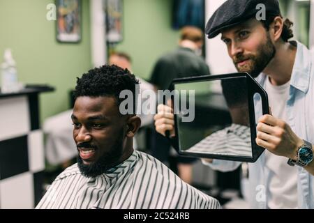 Porträt eines schönen schwarzen Mannes mit Kamm in den Haaren Blick in den Spiegel auf seine neue Frisur. Friseur zeigt dem Kunden seine Arbeit. Männliche Schönheit tre Stockfoto