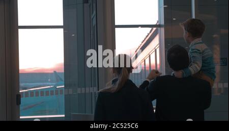 Mutter, Vater und Sohn warten am Flughafen und schauen Flugzeuge durch das Fenster bei Sonnenuntergang an. Sie zeigen auf etwas in der Ferne, Junge sitti Stockfoto