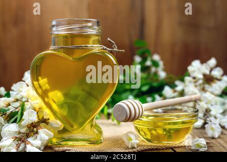 Süßer Honig in einem herzförmigen Glas und in einer Schüssel mit einem hölzernen Dipper und Akazienblumen auf dem alten Tisch. Selektiver Fokus? Stockfoto