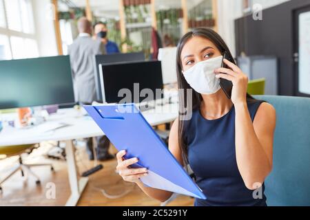 Junge asiatische Geschäftsfrau mit Gesichtsmaske wegen Covid-19 spricht im Büro am Handy Stockfoto