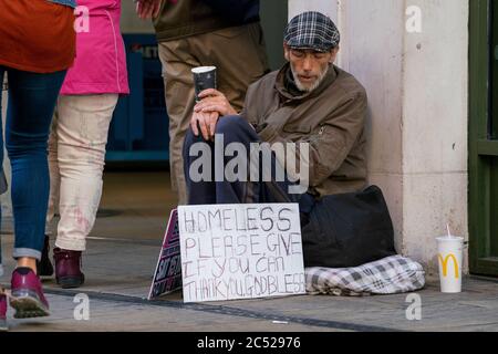 Ein Obdachloser sitzt draußen, mit einem McDonalds Kaffee in den Händen. Mit einem Schild, das um Geld bittet. Wie die Leute vorbei gehen, ohne sich zu kümmern. Stockfoto