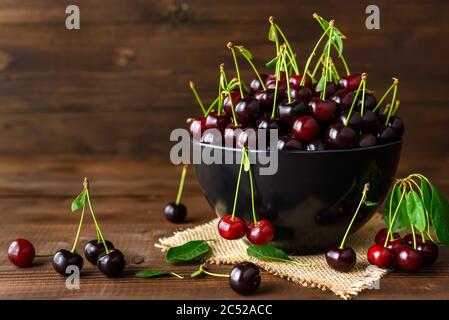 Frische Sauerkirschen in schwarzer Schale und grüne Blätter auf braunem Holztisch mit Platz für Text. Frische reife Sauerkirschen. Stockfoto