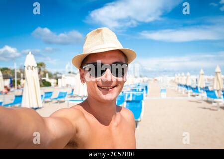 Lächelnder junger Mann mit Hut und Sonnenbrille, der Selfie mit dem Smartphone am Strand nimmt - lächelnder Teenager mit dem Smartphone, der Selfie nimmt Stockfoto