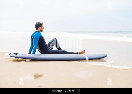 Junger Surfer im Neoprenanzug, der am Strand neben seinem Surfbrett sitzt, nach der morgendlichen Surfstunde. Stockfoto