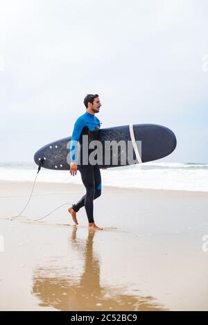 Junger Surfer im Neoprenanzug, hält Surfbrett unter seinem Arm und läuft am Strand nach der morgendlichen Surfstunde. Stockfoto