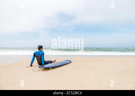 Junger Surfer im Neoprenanzug, der am Strand neben seinem Surfbrett sitzt, nach der morgendlichen Surfstunde. Stockfoto