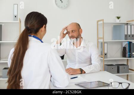 Reifer Mann mit Kopfschmerzen Beratung mit Ärztin in der Klinik. Medizinische Fachkraft hilft Patienten bei der Behandlung von Migräne Stockfoto