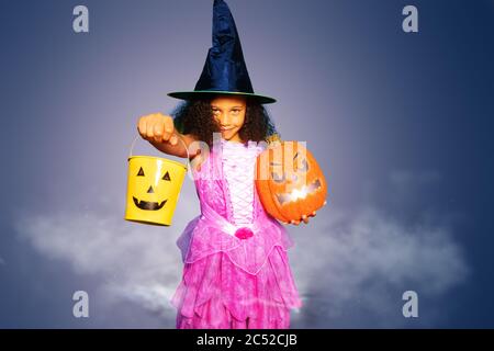 Portrait eines kleinen lockigen Haarmädchens im Hexenhut im Halloween Kleid halten gelben Bonbon Eimer und Kürbis über lila Hintergrund Stockfoto