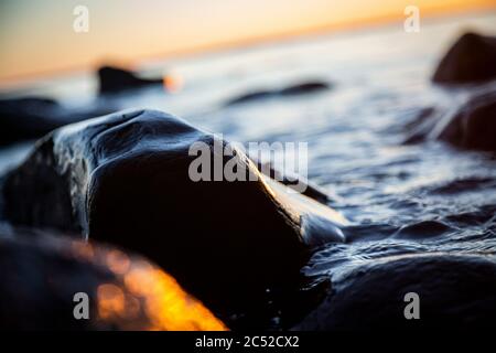 Nahaufnahme von Meereswellen, die auf einer nassen Küste brechen Steine Sommer Sonnenuntergang, Wasserspritzer, Sonnenstrahlen, Reflexionen auf Wassertropfen. Stockfoto