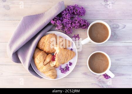 Zwei schöne Porzellanbecher Kaffee mit Milch mit Croissants mit Fliederblumen auf weißem Holztisch dekoriert. Perfektes Frühstückskonzept. Flach liegend. Stockfoto