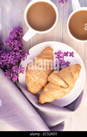 Zwei schöne Porzellanbecher Kaffee mit Milch mit Croissants mit Fliederblumen auf weißem Holztisch dekoriert. Perfektes Frühstückskonzept. Flach liegend. Stockfoto