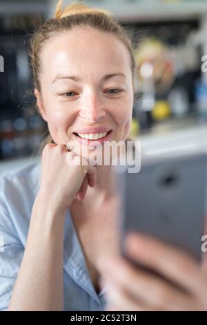 Junge fröhliche fröhliche fröhliche Frau zuhause in der Küche mit Social-Media-Apps auf dem Handy zum Chatten und Stechen, die mit ihr verbunden sind Stockfoto