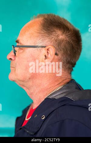 Ein Porträt eines älteren Mannes in blauem Mantel mit Hörgerät vor einem blauem Hintergrund. Stockfoto