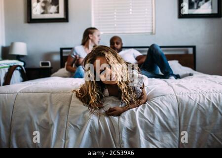 Junge Mädchen spielen auf dem Ende des Bettes mit Eltern im Hintergrund Stockfoto
