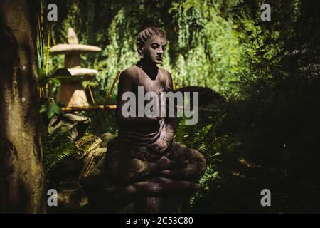 Stein sitzende Buddha-Statue von der Sonne im Schatten der Bäume im japanischen Garten während des sonnigen Sommertages mit Zen-Atmosphäre beleuchtet Stockfoto