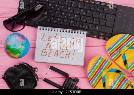 Gehen wir zum Strand auf Notebook mit Frau Reisenden Accessoires Brille Brieftasche und Flip-Flops auf rosa Tischplatte Hintergrund. Stockfoto