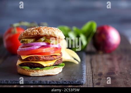 Vegetarischer Cheeseburger mit zwei Fleischersatzpasteten, Scheiben geschmolzenem Käse, Zwiebeln, Gurken, Salat und Tomaten auf einem frischen Sesamsamen BU Stockfoto