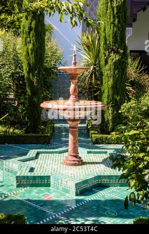 Ein brauner Marmorbrunnen in einer Gasse, die mit kleinen grünen Fliesen im üppigen Garten der Großen Moschee von Paris bedeckt ist. Stockfoto