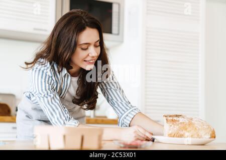 Bild von glücklichen schönen Frau trägt Schürze lächelnd während der Zubereitung von Brot in der modernen Küche Stockfoto