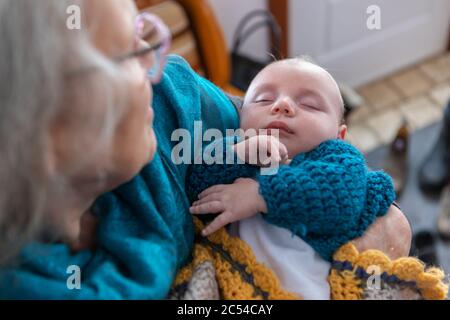 Selektive Fokusaufnahme eines kleinen Jungen, der in den Armen seiner Großmutter schläft. Aus der Fokussierung oben links schaut der stolze Senior auf das Kind Stockfoto