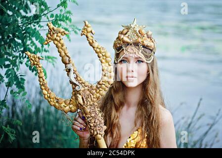 Fantasy Frau echte Meerjungfrau mit Dreizack Mythos Göttin des Meeres mit goldenen Schwanz sitzt in Sonnenuntergang auf Felsen.. Gold Haar Krone Muscheln Perlen Schmuck. Merma Stockfoto