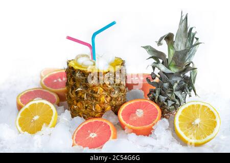 Fruchtcocktail in Ananas mit Saft und Stroh, steht auf Eis, daneben liegt eine Zitrone und eine Orange Stockfoto