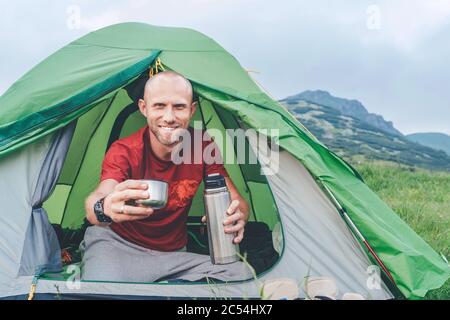 Lächelnder Mann im grünen Zelt, der den Tee aus einer Thermoskanne mit Berghintergrund trinkt. Er bietet dem Kameramann eine Teetasse an. Aktive Menschen genießen Stockfoto