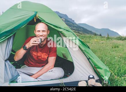 Mit freundlichen Grüßen lächelnder Mann im grünen Zelt, der den Kaffee aus einer Thermoskanne mit Berghintergrund trinkt. Aktive Menschen genießen Stockfoto