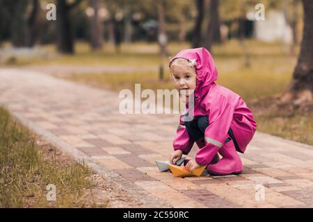 Hübsches Kind Mädchen 4-5 Jahre alt trägt Regenmantel und wasserdichte Stiefel spielen mit Papier Origami-Boot im Park im Freien. Blick auf die Kamera. Herbstsaison Stockfoto