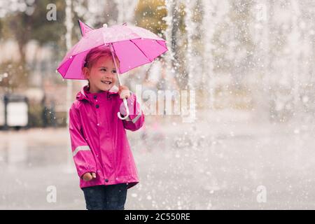 Lachende Kind Mädchen 4-5 Jahre alt hält rosa Regenschirm tragen Regenmantel über regen im Hintergrund Nahaufnahme im Park. Kindheit. Herbstsaison. Stockfoto