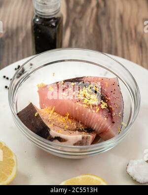 Zwei Stücke von frischen rohen Schwertfisch Steaks mariniert mit Salz, schwarzem Pfeffer und Zitronenschale in einer Schüssel auf Marmor Schneidebrett auf Holz Küchentisch, V Stockfoto