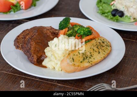 Köstliches geröstetes Rindfleisch mit Soße, Kartoffelpüree, Gemüse und Knoblauchbrotscheiben auf einem Teller Stockfoto