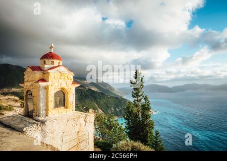 Kefalonia, Ionische Inseln, Griechenland. Bunte Proskinitari-Schrein-Laterne auf Sockel. Küstenlinie mit Wolkenlandschaft oben im Hintergrund. Stockfoto