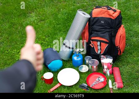 Bild von Campingwerkzeugen auf dem Gras - Rucksack, Zelt, Tank, Dosen, Kompass, etc., Tourist mit Daumen nach oben Stockfoto