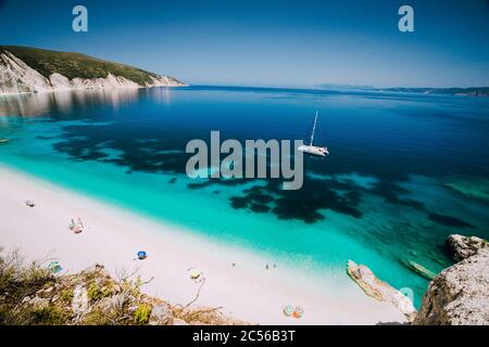 Weiße Katamaran-Yacht in klarem blauem Meerwasser. Touristen am Strand in der Nähe der azurblauen Lagune. Kefalonia, Griechenland. Stockfoto