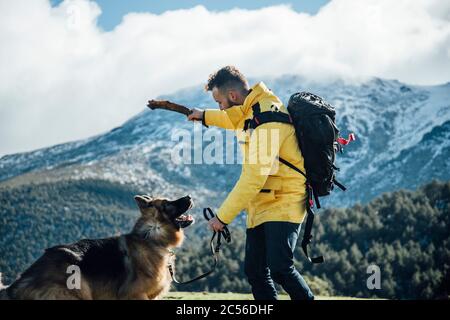 Junger Mann mit gelber Jacke und Rucksack spielt mit Schäferhund in den Bergen. Stockfoto