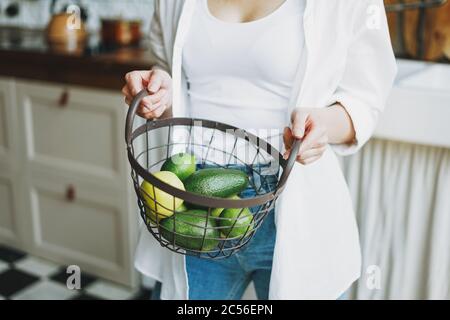 Crop Foto von jungen Frau in weißem Hemd halten Korb mit grünen Früchten und Gemüse in den Händen auf Küche Stockfoto